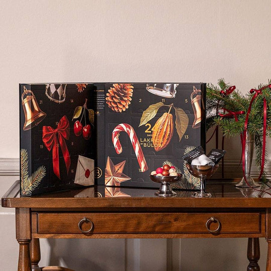 Chokladshopen Lakrids By Bülow adventskalender med svart och guldig juldesign på ett träbord med juldekorationer.