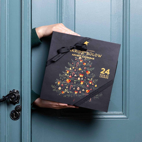 Chokladshopen Lakrids By Bülow adventskalender med julgransillustration i guld på svart bakgrund hållen i händerna
