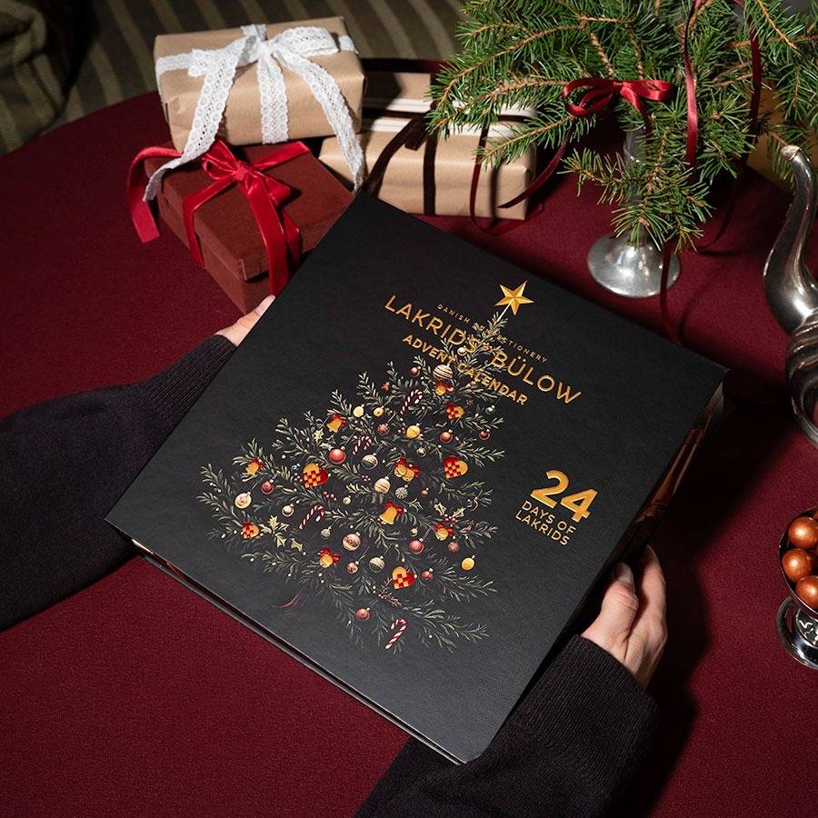 Chokladshopen Lakrids By Bülow adventskalender med svart design och guldillustration av en julgran på ett bord